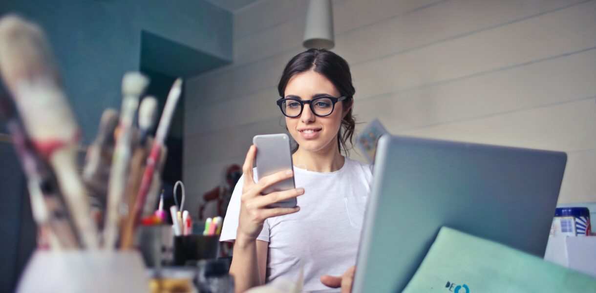 Young woman sitting in front of a computer and looking at her smartphone.