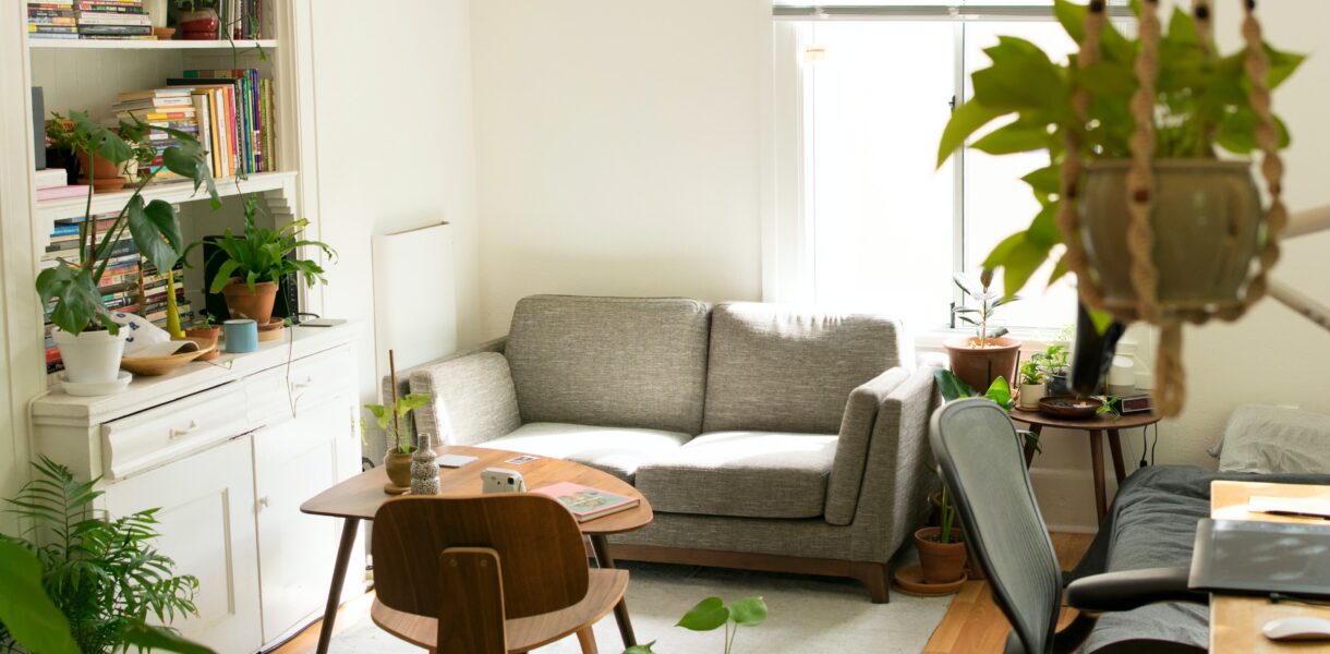 Room with a lot of green plants, a shelf with books, two tables and a bed.