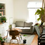 Room with a lot of green plants, a shelf with books, two tables and a bed.